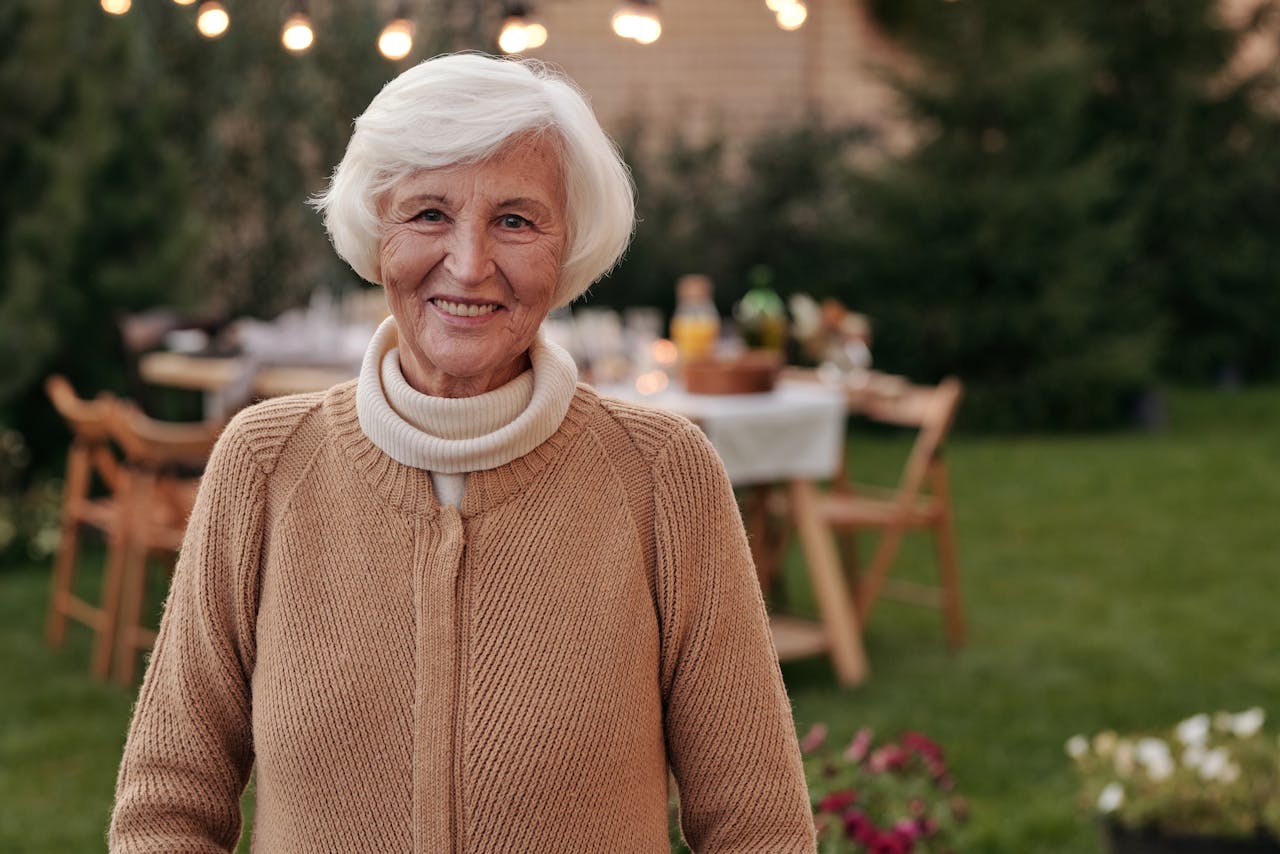 Senior woman in a tan sweater smiling in front of a table set for an outdoor family gathering.